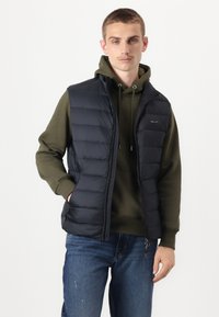Black padded vest with a zipped front, worn over an olive green hoodie. Features side pockets and a subtle logo on the chest. Denim jeans below.