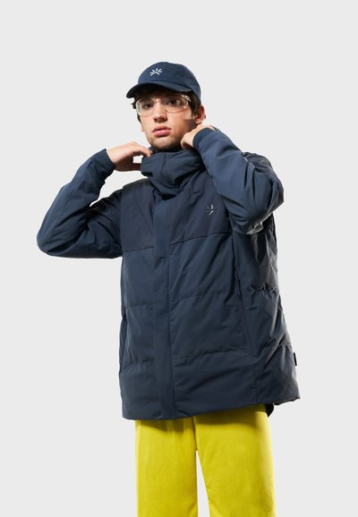 Marinblå pufferjacka med hög krage och dragkedjefickor, stylad med en matchande keps. Modellen bär ljusgula byxor och klara glasögon.