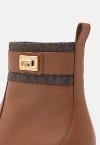 Bottine en cuir marron avec garniture en tissu à motif sombre et plaque logo MK dorée près de la languette supérieure.