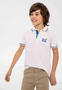 Bílá polo tričko s modrým límcem, obsahuje logo "SEA & SURF" v modré barvě. Krátké rukávy s vícebarevnými pruhy na manžetách. Béžové kalhoty.
