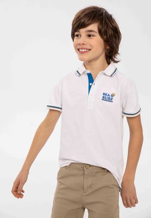 Biała koszulka polo z niebieskim kołnierzykiem, z niebieskim logo "SEA & SURF". Krótkie rękawy z wielokolorowymi paskami na mankietach. Beżowe spodnie.