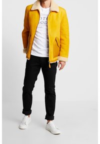 Gul shearling jakke med lynlås, sidelommer og en krave af kunstpels, parret med en hvid t-shirt, sorte jeans og hvide sneakers.