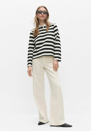 Femme portant un chemisier à manches longues rayé noir et blanc, un pantalon ivoire à jambes larges, des chaussures noires pointues et des lunettes de soleil noires sur fond blanc.