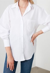 Camicia bianca con bottoni, tasca sul petto, maniche lunghe e vestibilità rilassata. Indossata con jeans blu.