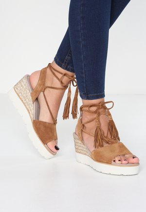 Sandales compensées en daim marron avec liens à nouer à la cheville et pompons, portées avec un jean skinny bleu foncé sur fond blanc.
