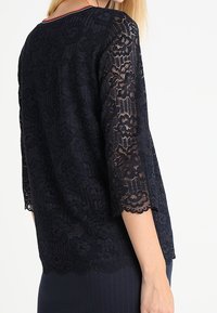 Top in pizzo blu navy con maniche a 3/4, orlo scallop e motivo floreale. Presenta un collo rotondo con dettagli in rosso.