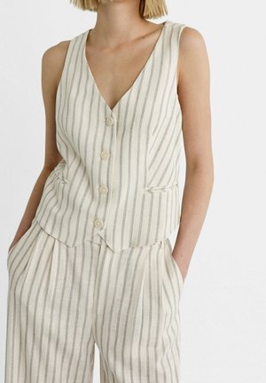 Vrouw met een mouwloze beige gilet en bijpassende hooggelegen gestreepte broek, met handen in de zakken tegen een effen achtergrond.