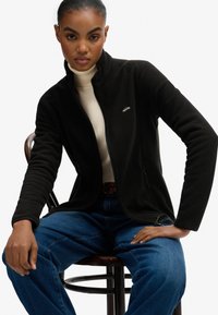 Zwart fleecejack met hoge kraag, ritssluiting aan de voorkant en zijzakken, gecombineerd met een crèmekleurige coltrui en blauwe spijkerbroek.