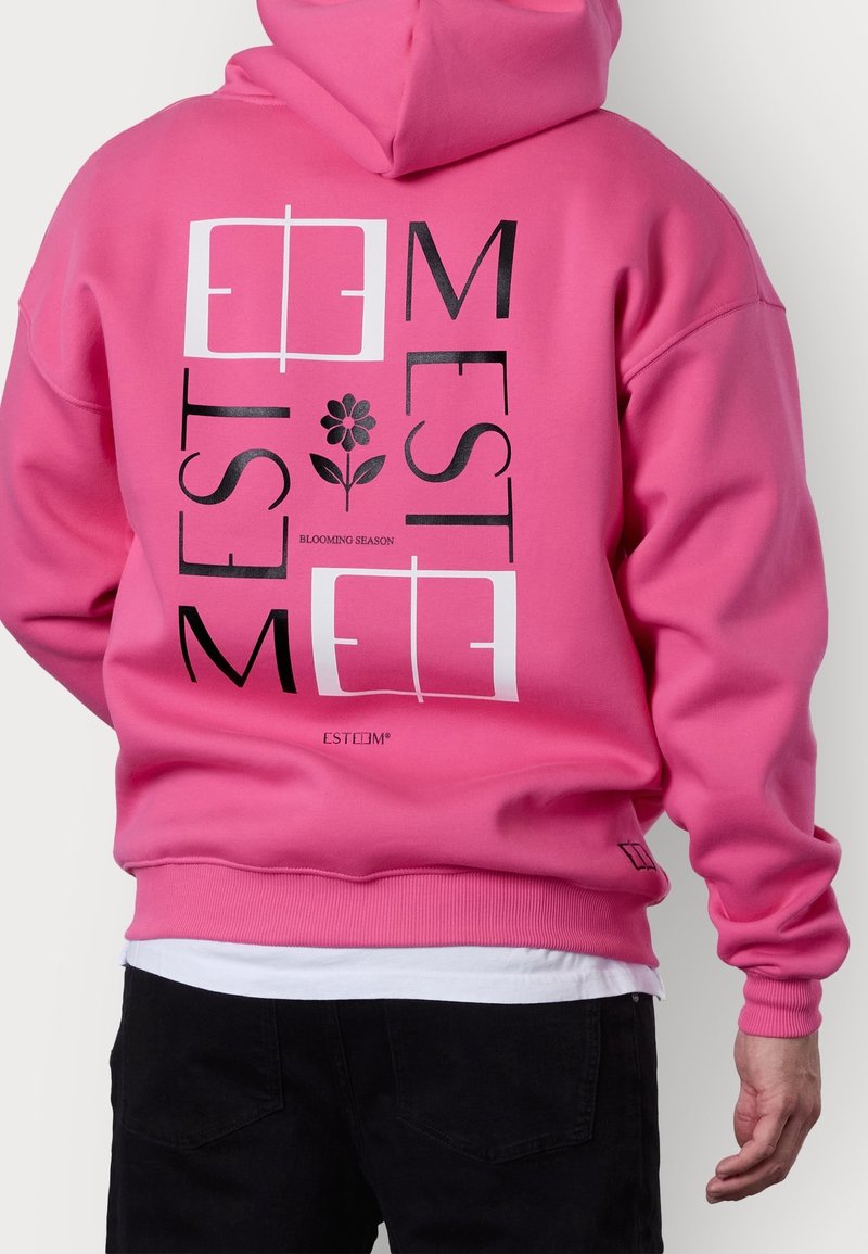 Sweat à capuche rose vif avec texte graphique noir et blanc « ESTEEM » et une illustration de fleur au dos, porté avec une chemise blanche et un pantalon noir.