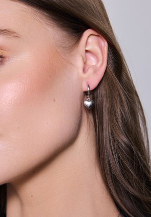 Srebrne kolczyki w kształcie kół z małym zawieszką w formie serca, zaprezentowane na uchu modelki, ukazujące błyszczące wykończenie i elegancki design.