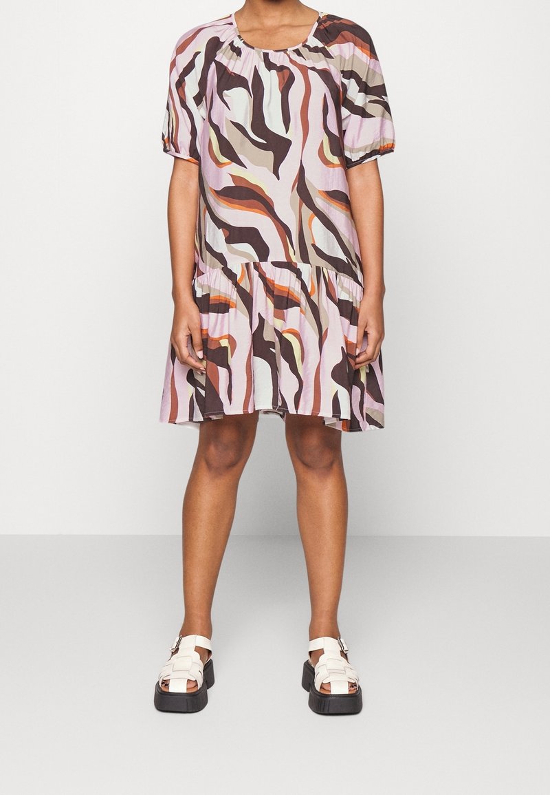 Kortärmad klänning i ett rosa och brunt vågigt mönster, med en lager på lager kjol och en rund halsringning. Kombinerad med vita klumpiga sandaler.