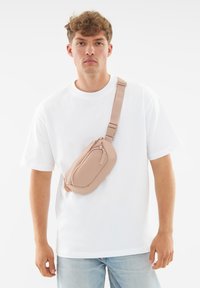 Marsupio rosa chiaro realizzato in materiale sintetico liscio, dotato di chiusura con zip e tracolla regolabile, indossato sopra una T-shirt bianca.