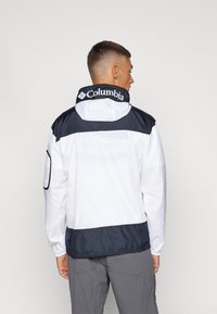 Jachetă de vânt albă și bleumarin cu glugă, având branding Columbia pe guler și manșete elastice, din material sintetic neted.