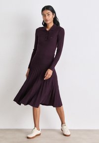 Uska pripijena haljina s rebrastim uzorkom u dubokoj ljubičastoj boji, s ovratnikom, zatvaranjem na gumbe i nabranim suknjom. Uparena s bijelim tenisicama s smeškastim đonovima.