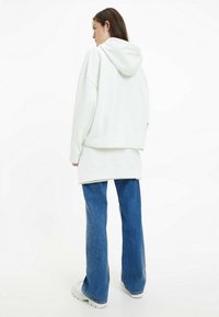 Felpa bianca oversized con cappuccio, in tessuto testurizzato, indossata sopra un top lungo, abbinata a jeans blu a zampa d'elefante e scarpe bianche.