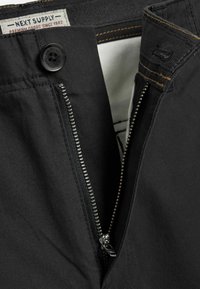 Schwarze Hose mit einem Metallreißverschluss, Kunststoffknopfverschluss und kontrastierenden Nähten. Innenfutter sichtbar am Bund.