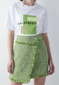 Bílý tričko s zeleným grafickým designem a černým textem. Zelená obručová sukni s třásněmi z texturovaného materiálu s vzorem malých tkanin.