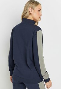Felpa blu navy con colletto alto, featuring pannelli laterali grigi e polsi a coste. Tessuto liscio con un design a vestibilità rilassata.
