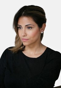 Una camicia nera a maniche lunghe, in tessuto liscio. Capelli lunghi e lisci con morbide onde. Orecchini a perla rotondi. Sfondo neutro. Focus sul volto e sull'espressione.