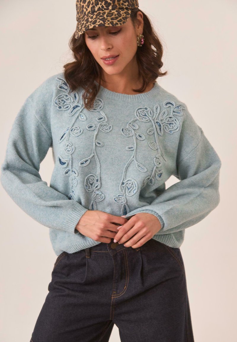 Suéter de punto azul claro con bordado floral y cuentas en el frente; corte relajado con puños y dobladillo de canalé; combinado con jeans azules.