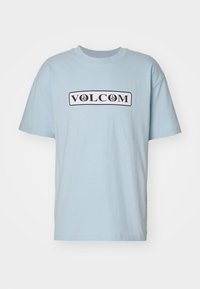 Camiseta de algodón azul claro con un logo negro de "VOLCOM" sobre un fondo rectangular blanco. Diseño de manga corta y cuello redondo.