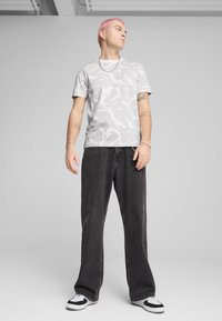 Grå mønstret t-shirt med hvide abstrakte designs, parret med sorte brede jeans og sorte-hvide sneakers. Afslappet streetwear stil.