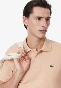 Camiseta polo de melocotón claro con un acabado texturizado, tres botones y un pequeño logo de cocodrilo verde en el lado izquierdo del pecho. Material de algodón.