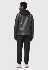 Chaqueta de cuero negra con capucha, que presenta una textura suave y un corte ajustado. Llevada sobre pantalones jogger negros y zapatillas ligeras.