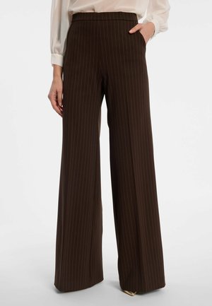 Vrouw draagt hooggetailleerde, donkerbruine wijde broek met dunne verticale strepen en een ingestoken wit bloesje, één hand in de zak.