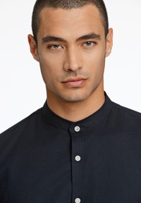 Cămașă de culoare albastru marin cu guler mandarin, prevăzută cu o placket cu nasturi de culoare deschisă. Material neted și croială ajustată.