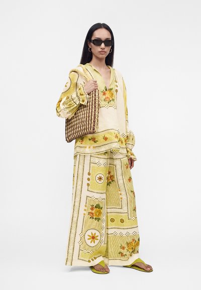 Γυναίκα με μακριά σκουρόχρωμα μαλλιά που φοράει γυαλιά ηλίου, φαρδιά μπλούζα με λουλουδάτο σχέδιο και ταιριαστό παντελόνι φαρδιάς γραμμής, κρατώντας πλεκτή τσάντα, όρθια.