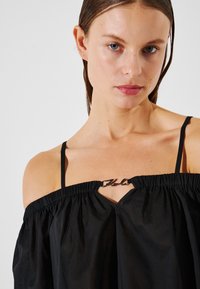 Top nero a spalla scoperta realizzato in tessuto leggero con scollatura arricciata e spalline sottili, caratterizzato da un accento argentato sul petto.