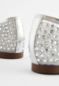 Ballerines argentées avec semelles marron, dotées de panneaux en mesh décorés de multiples strass brillants, vues de dos en gros plan.
