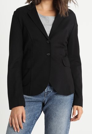 Žena v priléhavém černém saku přes šedé tričko, v modrých džínách, na jednobarevném bílém pozadí.