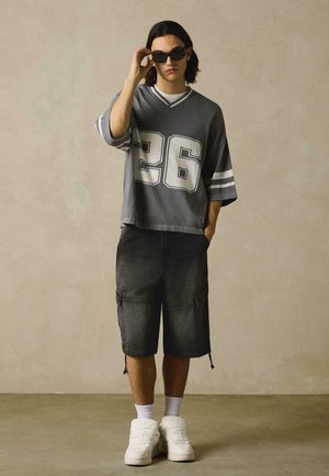 Bărbat tânăr purtând ochelari de soare negri, tricou sport larg gri cu numărul 26, pantaloni scurți cargo negri, șosete albe și adidași albi.