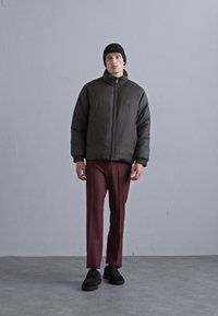 Jachetă puffer de culoare verde închis, cu guler înalt și închidere cu fermoar, combinată cu pantaloni burgund și pantofi negri, pe un fundal neutru.