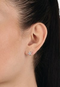 Pendientes en forma de estrella plateada con una gema central brillante, presentando superficies suaves y un acabado pulido, sujetos de forma segura al lóbulo de la oreja.