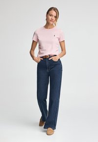Rosa Kurzarm-T-Shirt, blaue hoch geschnittene Jeans und helle Slipper. Das T-Shirt hat ein kleines Logo. Glatte Textur.