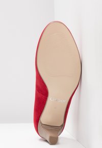 Chaussure à talon haut en daim rouge inclinée vers le haut, montrant une semelle beige lisse avec le nom de marque « Gabor » et la taille 4, sur fond blanc.