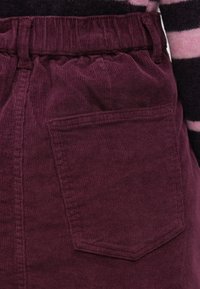 Bordó kordbársony szoknya gyűrt derékrésszel és egyetlen hátsó zsebbel. Függőleges bordázott textúrával és tiszta szegéllyel rendelkezik.