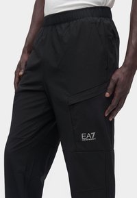 Άνδρας που φοράει μαύρο παντελόνι EA7 Emporio Armani με ελαστική μέση και πλευρική τσέπη, μερικώς ορατό λευκό πουκάμισο.