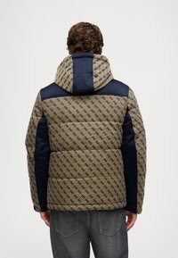 Homem em um casaco acolchoado com padrão bege e azul-marinho, com capuz, de costas, combinado com jeans pretos desbotados.