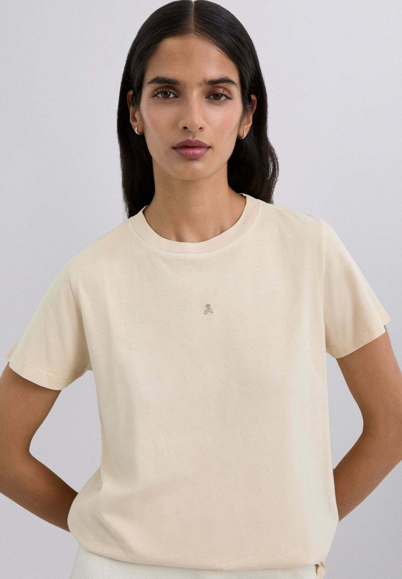 Camiseta de algodón beige con cuello redondo y mangas cortas, que presenta un pequeño logo de calavera en un color sutil en el pecho.