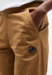 Κοντινή λήψη ενός ατόμου που φοράει καφέ παντελόνι με ελαστική μέση, χέρι στην τσέπη και μαύρο λογότυπο μαμούθ κοντά στην τσέπη.