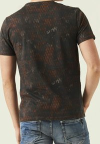 Muž v tmavém, vzorovaném tričku s krátkým rukávem a modrých džínech, viděný zezadu na jednobarevném pozadí.