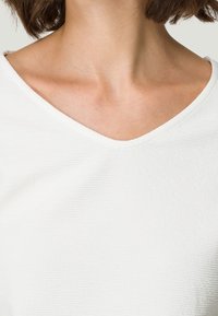 Cuello y parte superior del pecho de una mujer visibles, lleva una camiseta blanca texturizada con cuello en V y cabello corto castaño.