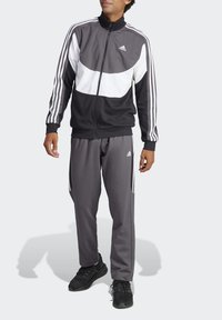 Tuta sportiva nera e grigia con giacca con zip e dettagli bianchi, tre strisce bianche sulle maniche e pantaloni abbinati.
