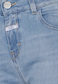 Jean en denim bleu clair montrant la taille avant, une passante de ceinture, un bouton marqué "CLOSED" et une petite étiquette avec le nom de la marque cousue près de la poche.