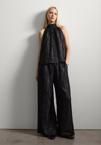 Zwarte, gekreukelde stoffen outfit met een mouwloze top met een hoge hals en wijde broekspijpen, beide met een gestructureerd oppervlak.