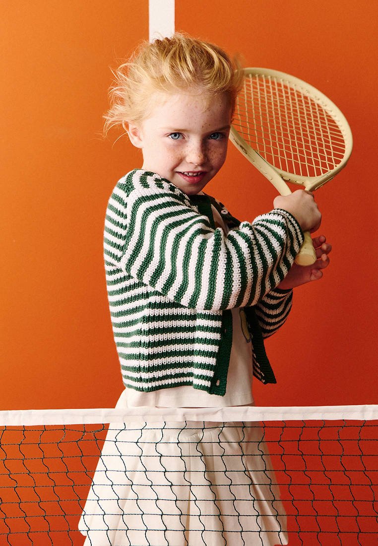 Jeune fille aux cheveux roux et pull rayé tenant une raquette de tennis derrière un filet devant un mur orange.